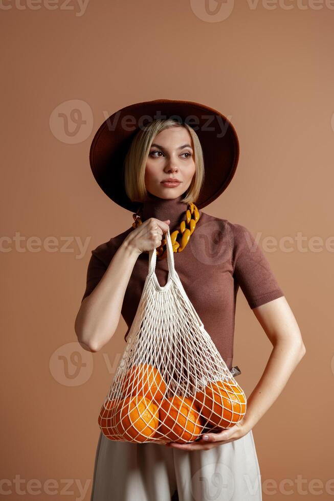 Fashionable woman in elegant hat carrying mesh bag with oranges against brown background photo