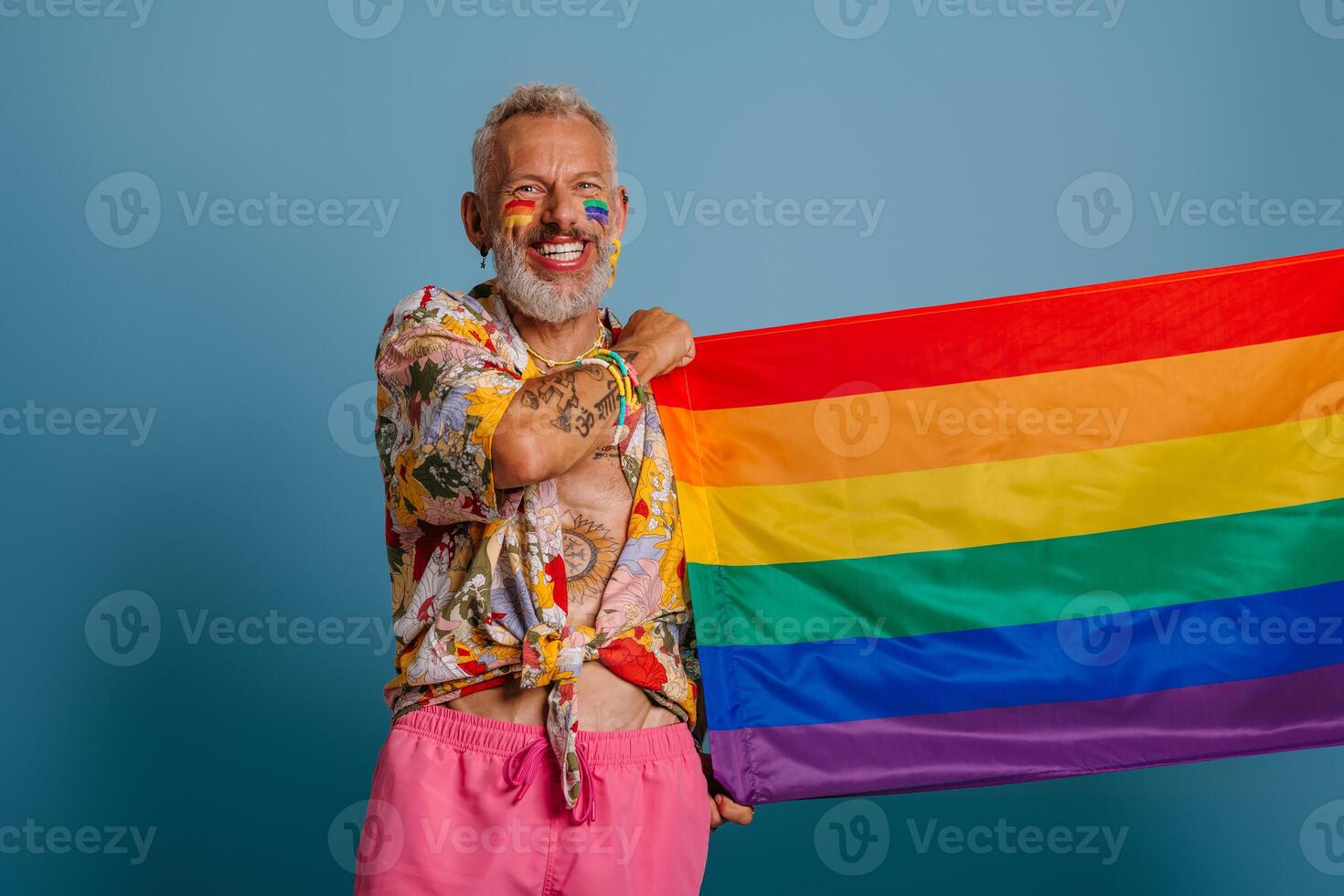 Cheerful mature gay man carrying rainbow flag and smiling while standing against blue background photo