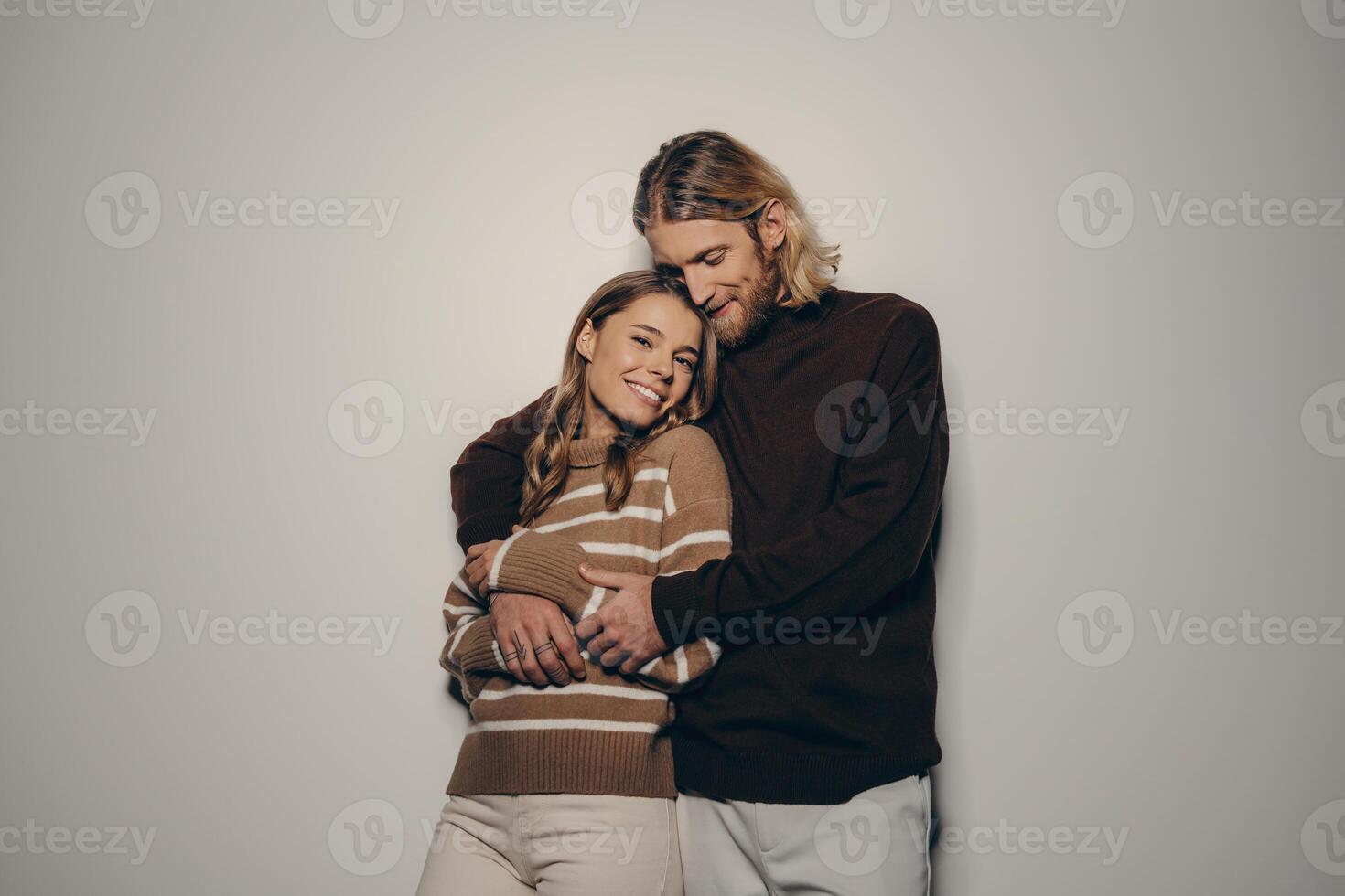 Fashionable young loving couple in cozy warm wear embracing while standing on beige background photo