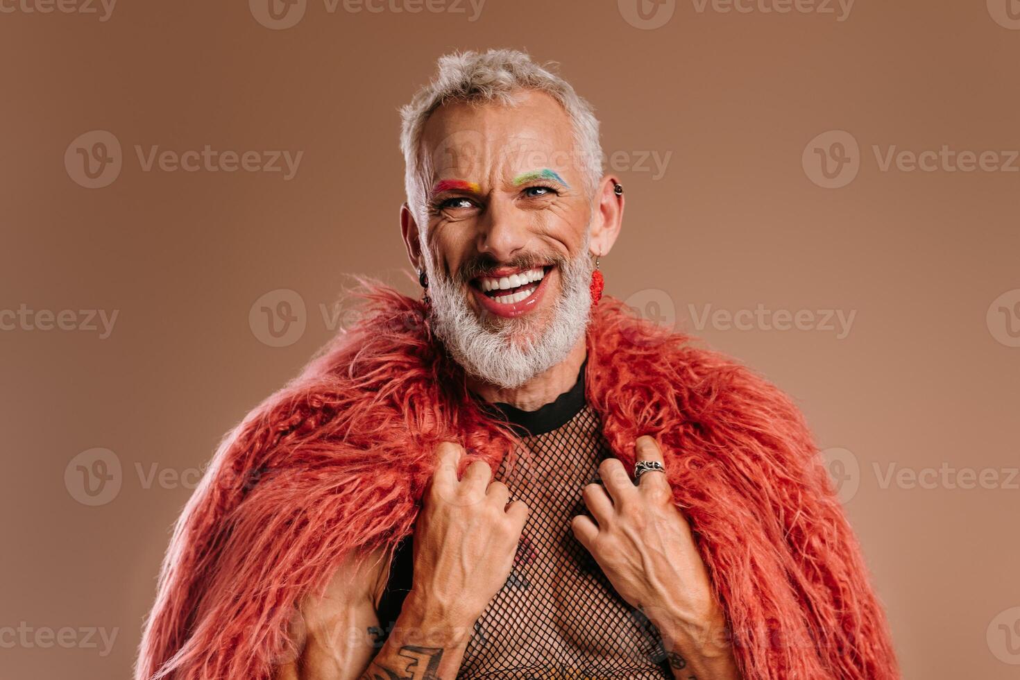 Happy mature gay man with rainbow colored eyebrows wearing fluffy coat against brown background photo