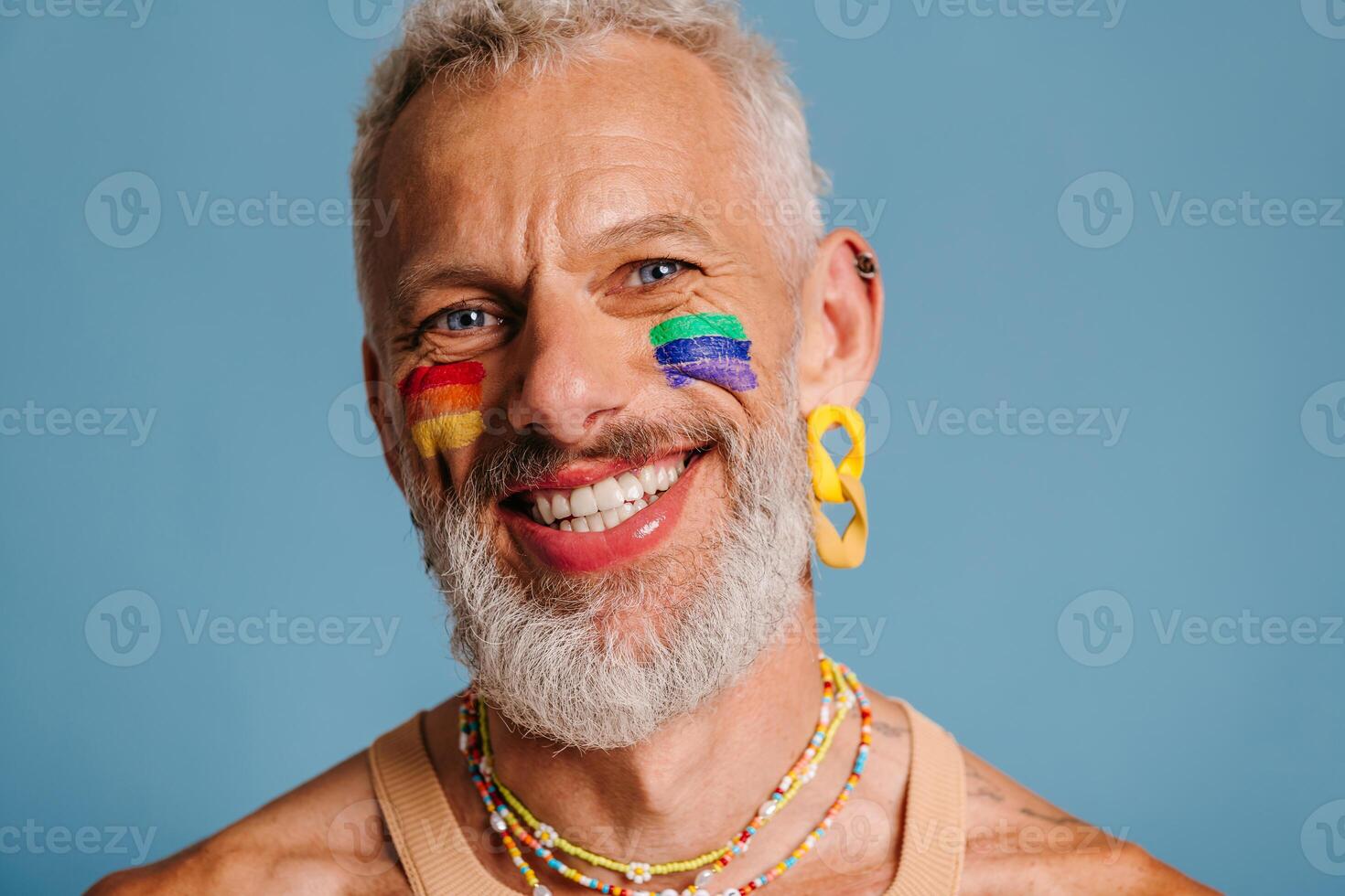 Portrait of mature gay man with rainbow flag make-up looking at camera and smiling against blue background photo