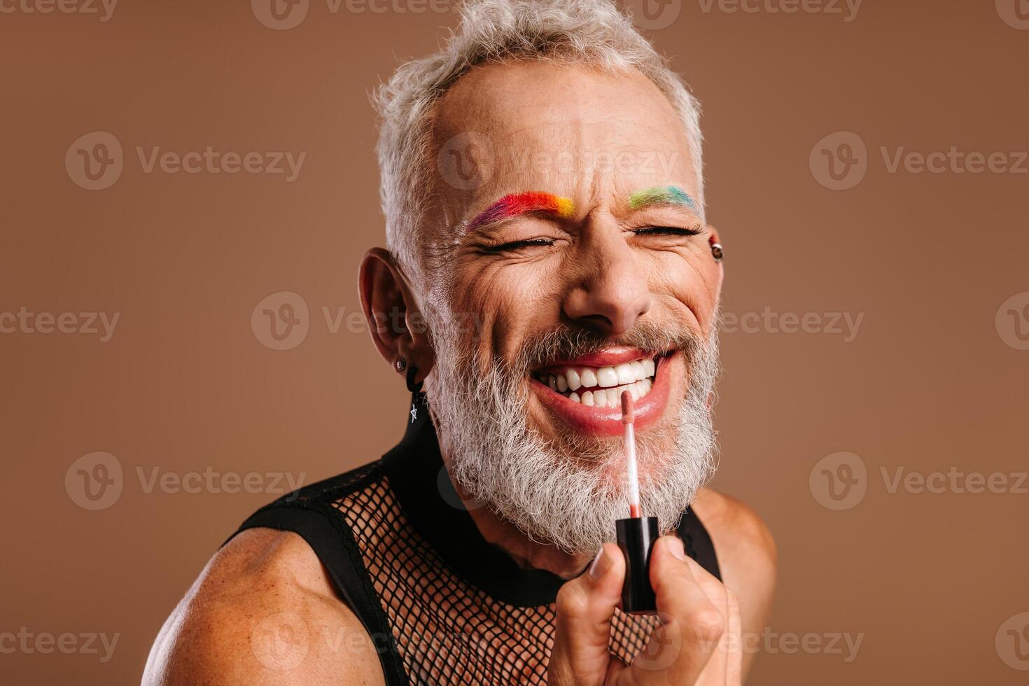 Happy mature gay man with sincere smile applying make-up against brown background photo