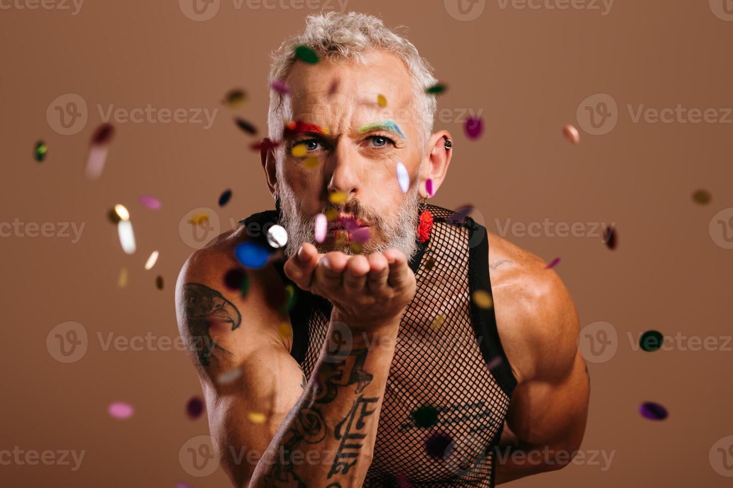 Bearded mature gay man with stage make-up blowing colorful confetti against brown background photo