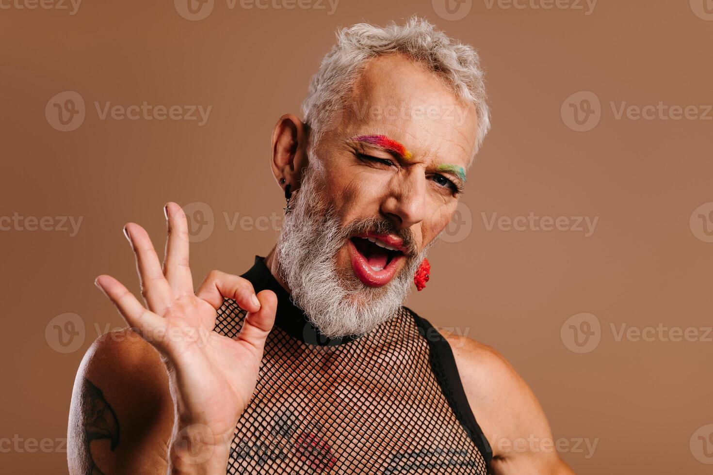 Playful mature gay man with rainbow colored eyebrows gesturing and winking against brown background photo