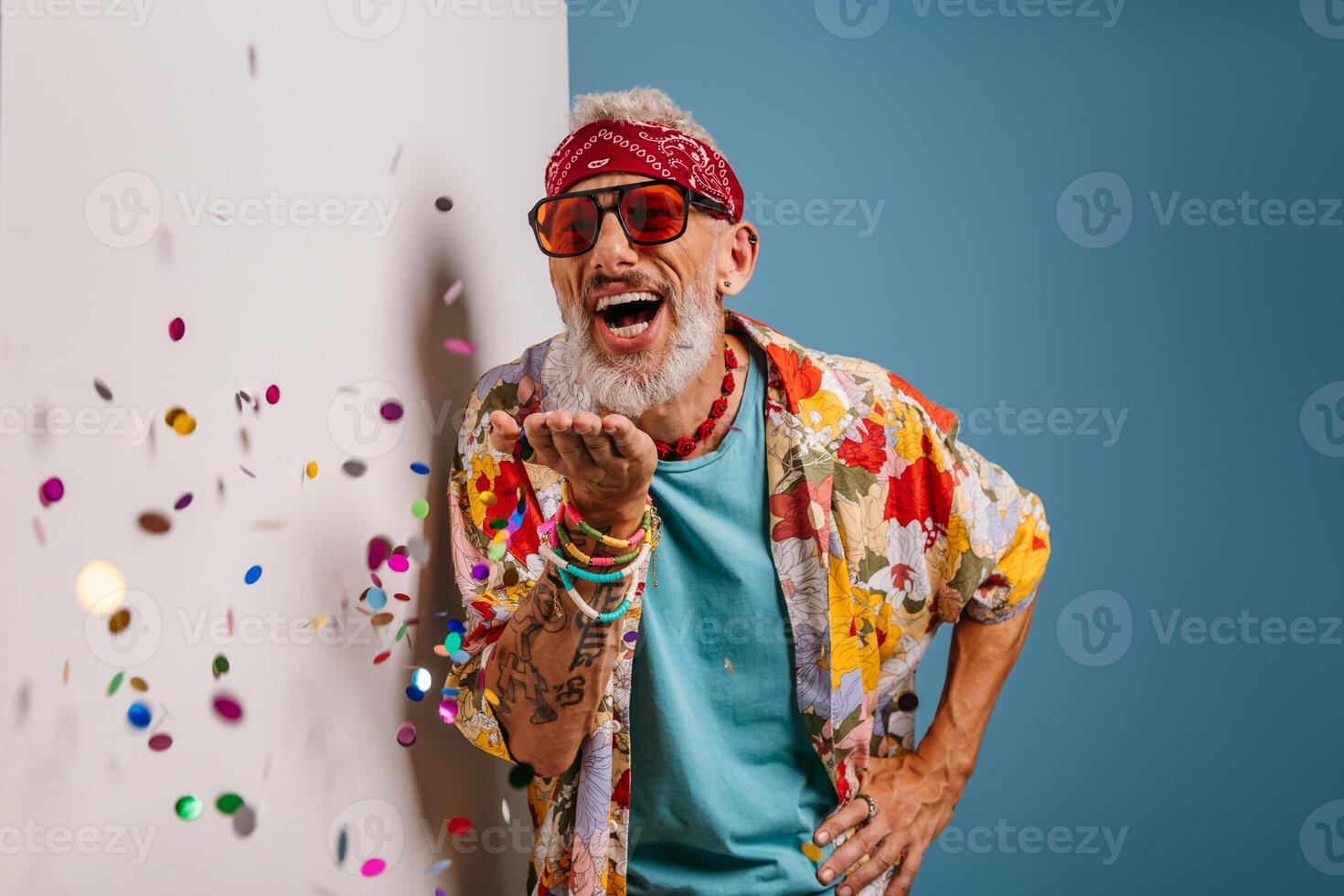 Happy senior man in funky shirt and bandana blowing colorful confetti on white and blue background photo
