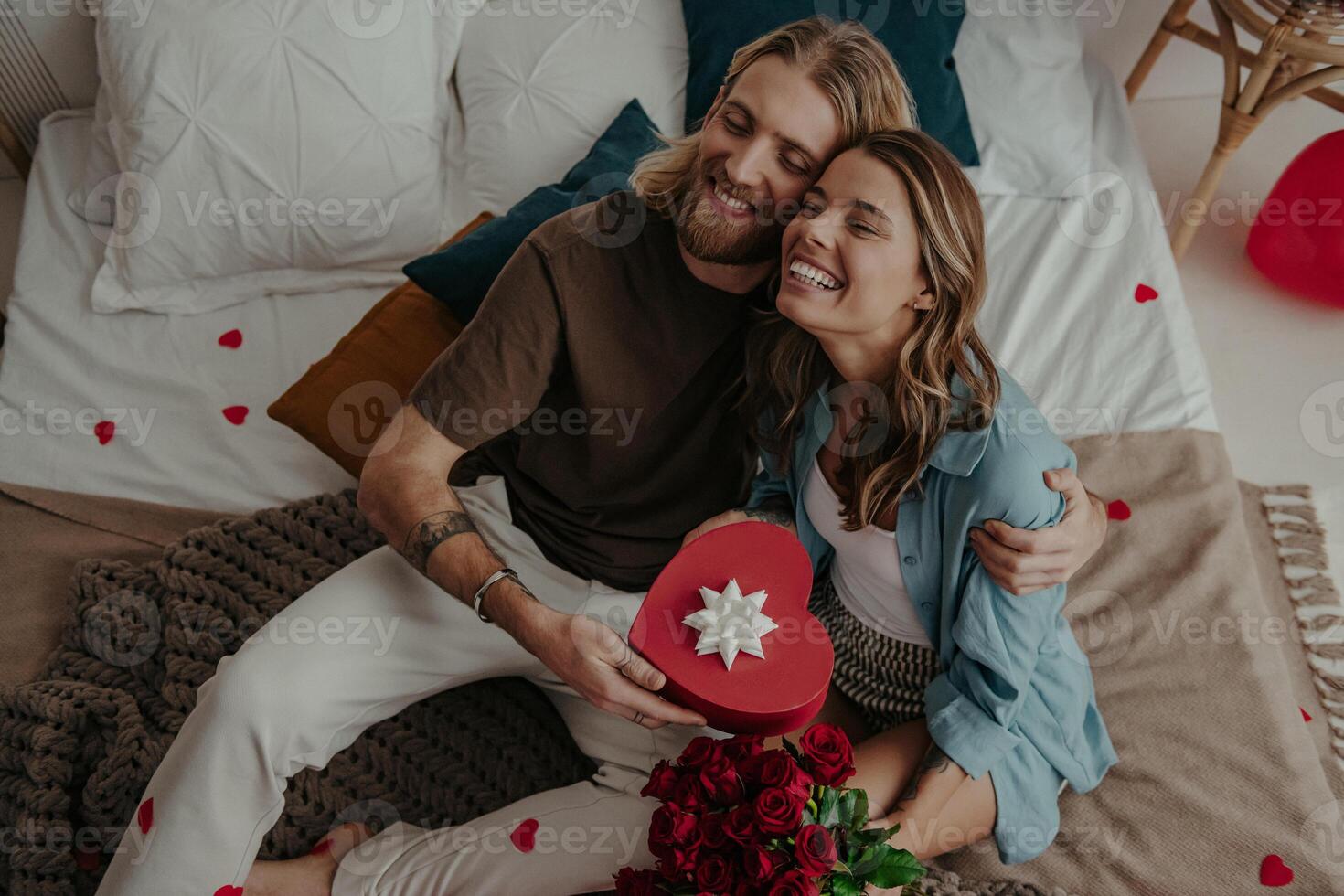 Top view of loving couple embracing and holding heart shape gift box while sitting on bed at home together photo