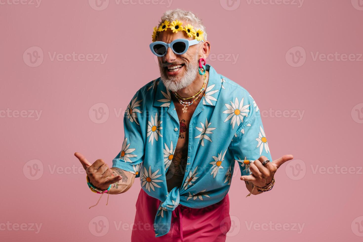 Joyful senior man in stylish shirt and floral wreath on head gesturing against pink background photo
