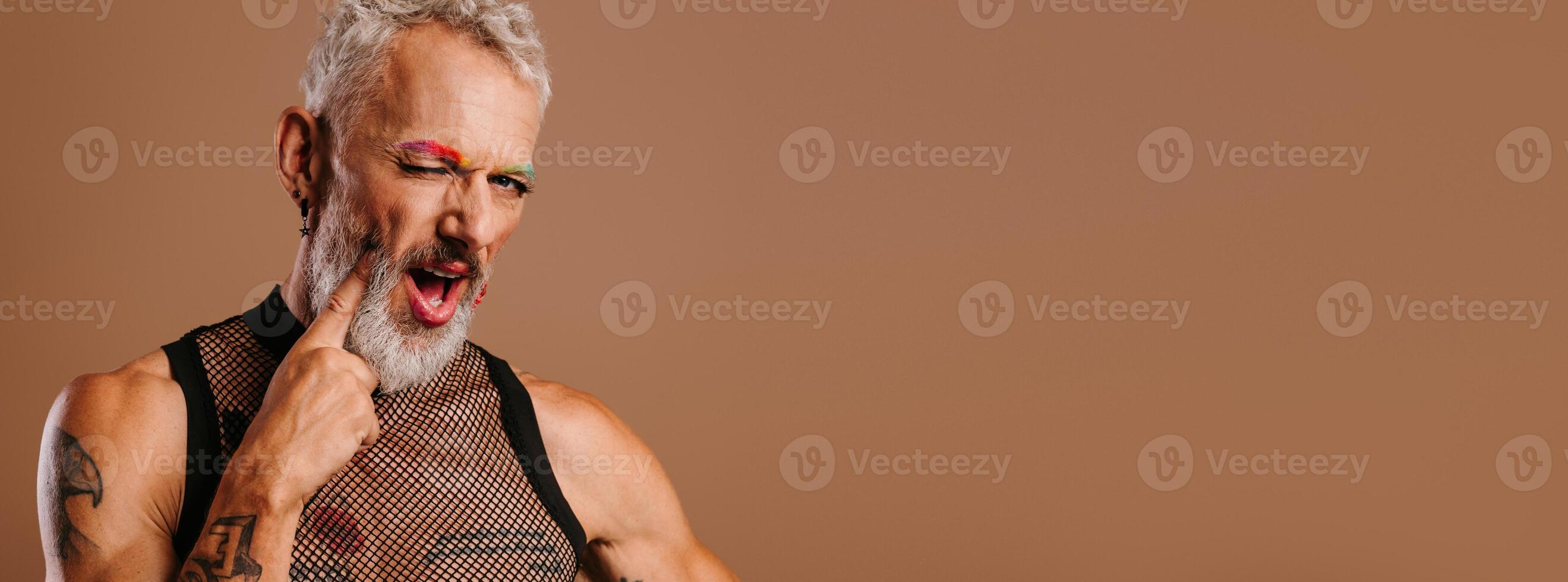 Playful mature gay man with rainbow colored eyebrows touching face with finger against brown background photo