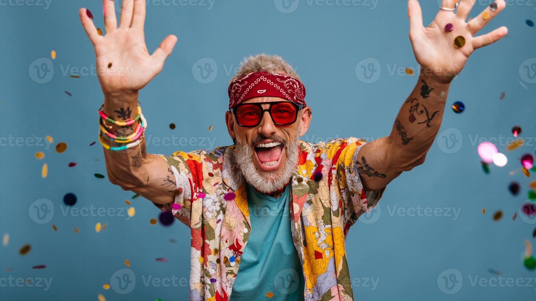 Joyful senior man in funky shirt and bandana throwing colorful confetti on blue background photo