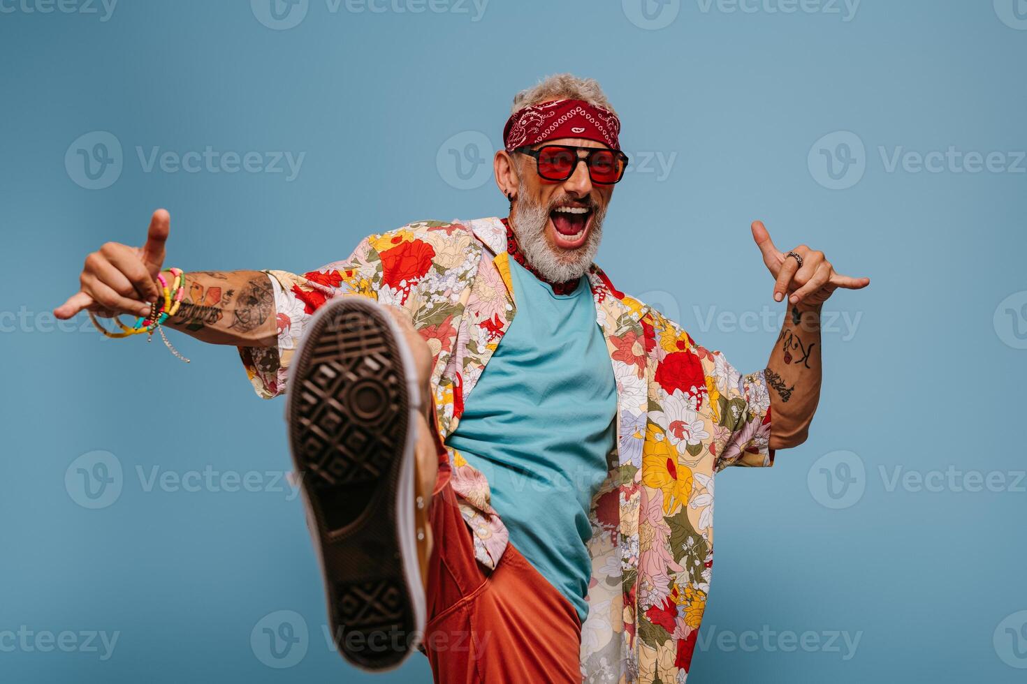 Cool hipster senior man in stylish funky shirt kicking leg and gesturing against blue background photo