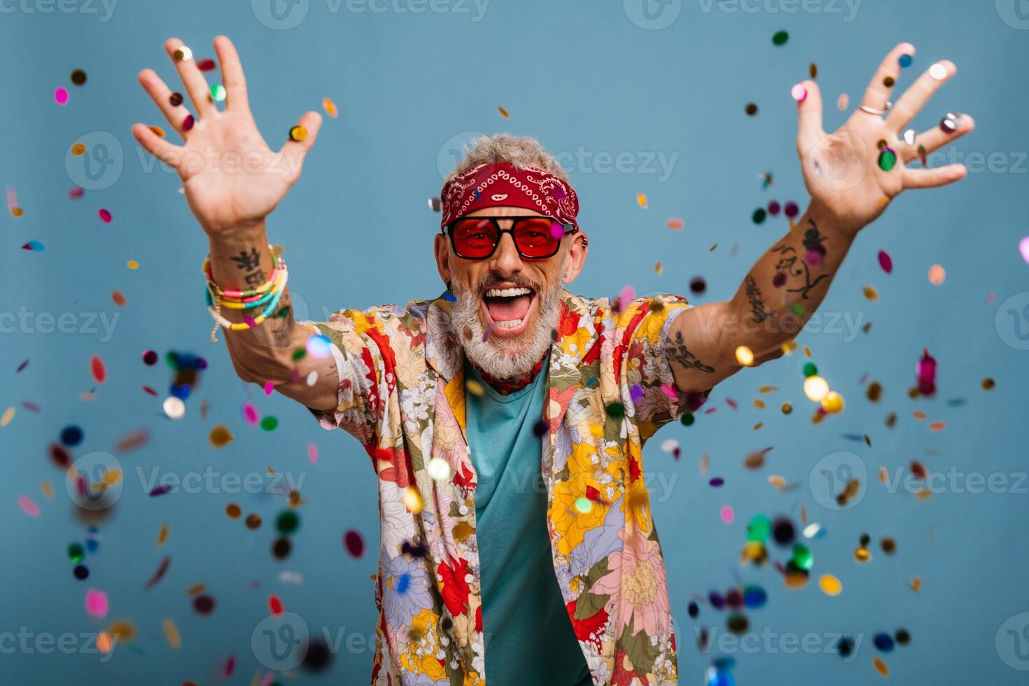 Happy senior man in funky shirt and bandana throwing colorful confetti on blue background photo