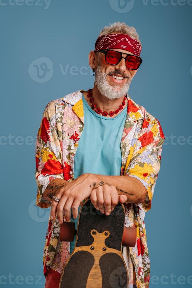 Hipster senior man in stylish funky shirt leaning on longboard and smiling against blue background photo
