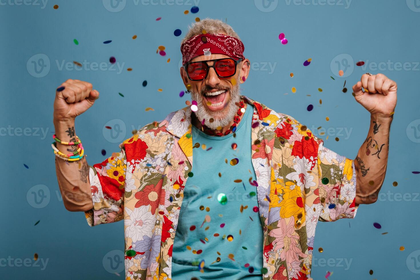 Happy senior man in funky shirt and bandana gesturing while confetti flying around on blue background photo
