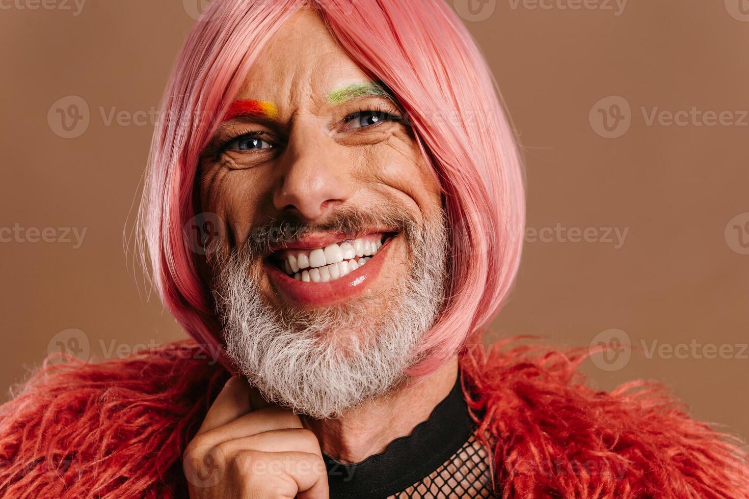 Portrait of mature gay man with rainbow colored eyebrows wearing pink wig against brown background photo