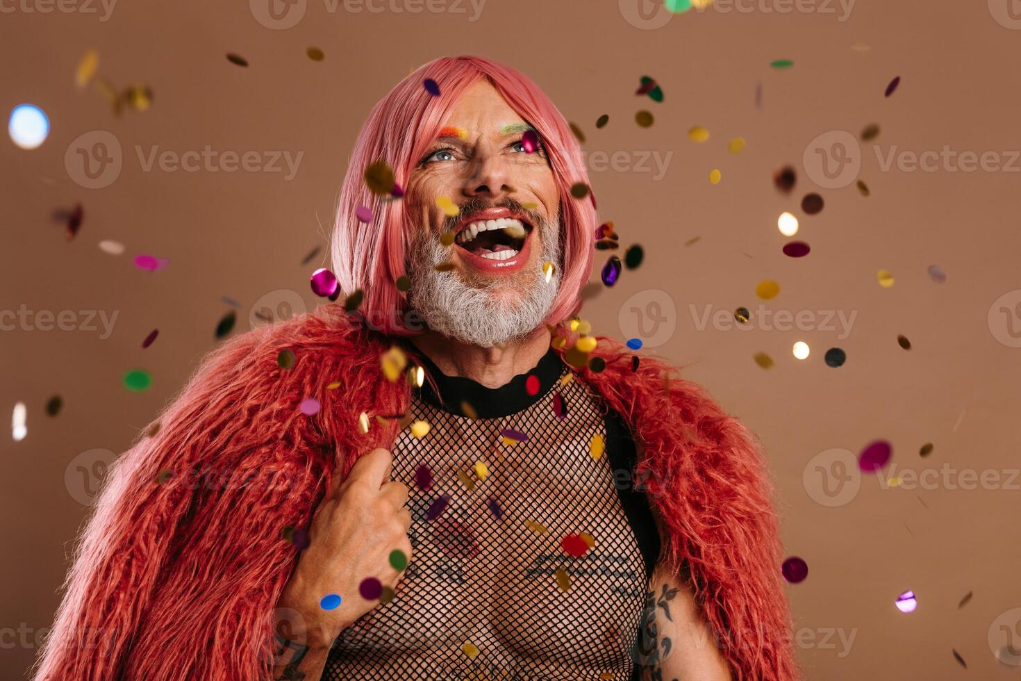 Happy gay man in pink wig looking at the colorful confetti flying around against brown background photo