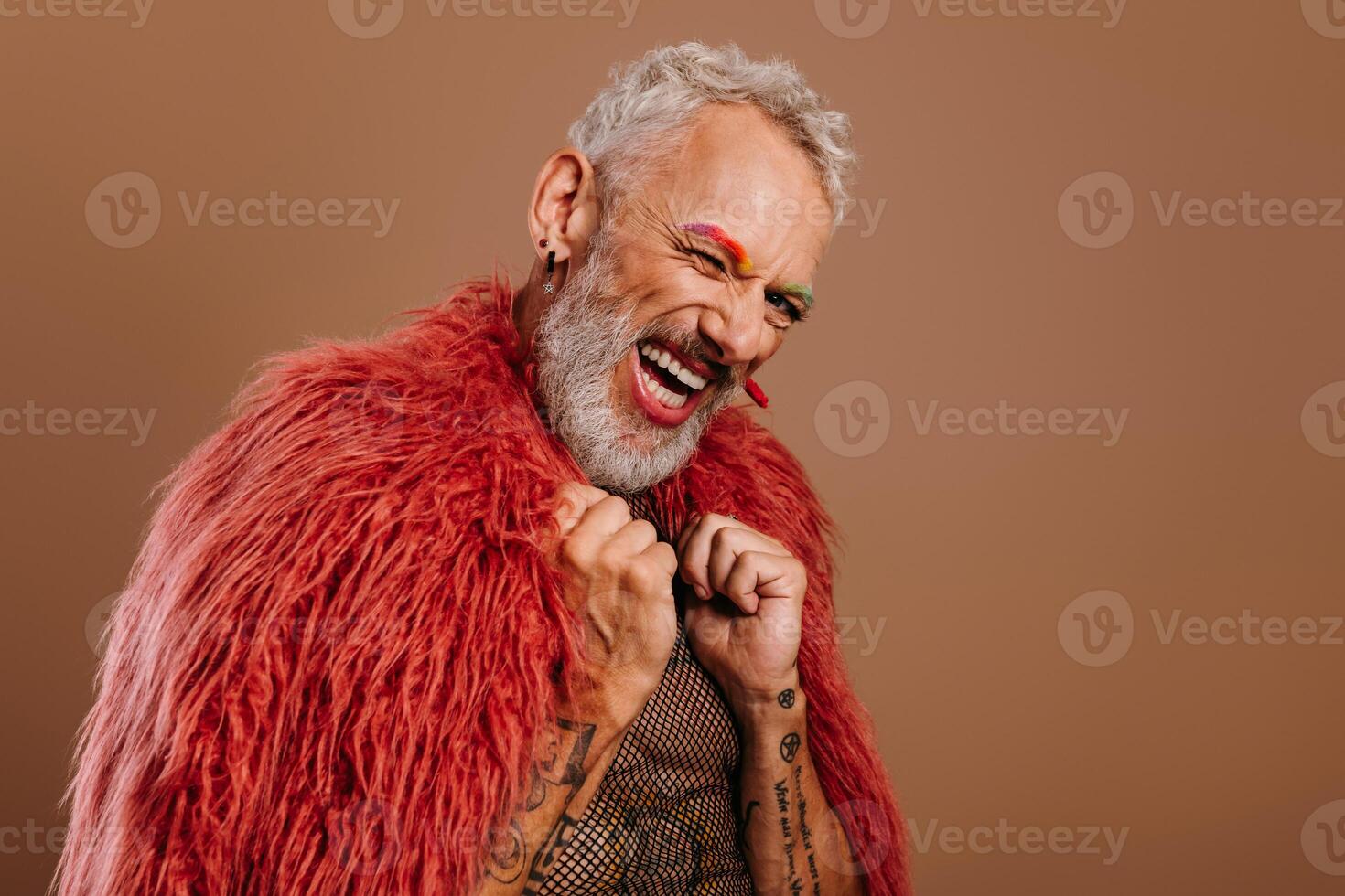 Playful mature gay man with rainbow colored eyebrows wearing fluffy coat against brown background photo