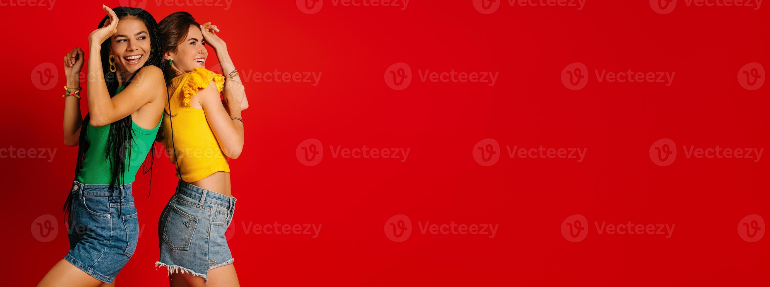Two attractive young women in colorful wear standing back to back while dancing against red background photo