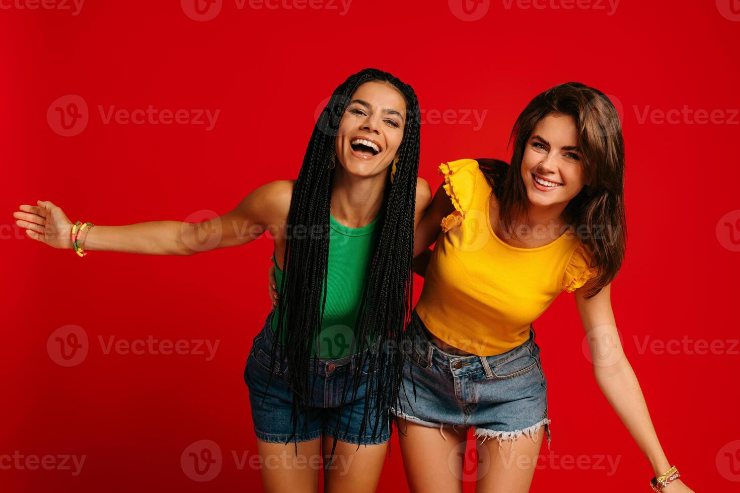Two beautiful young women in colorful wear embracing and smiling against red background photo