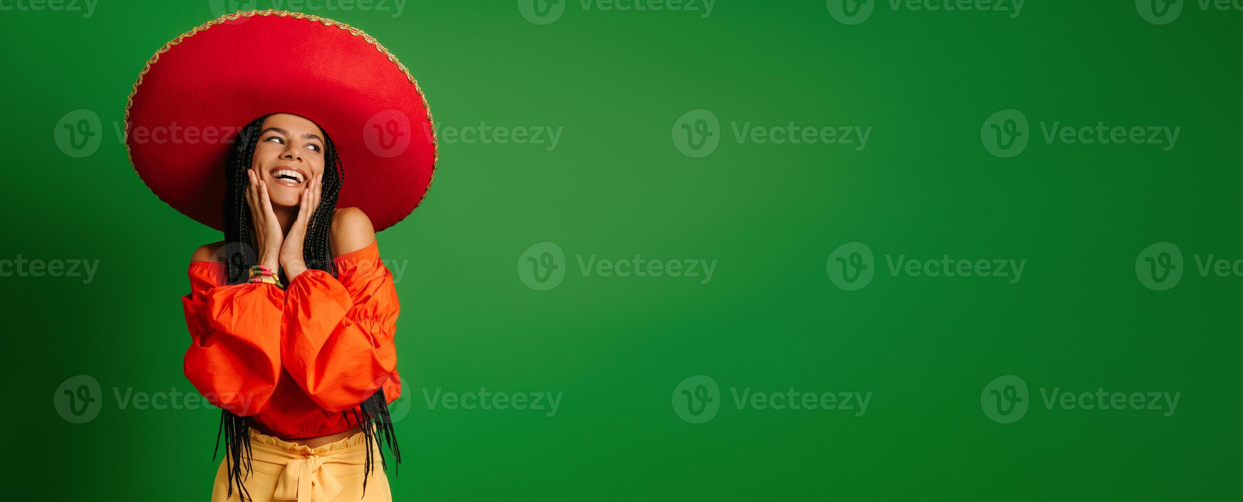 Happy young Mexican woman in Sombrero touching face and smiling against green background photo