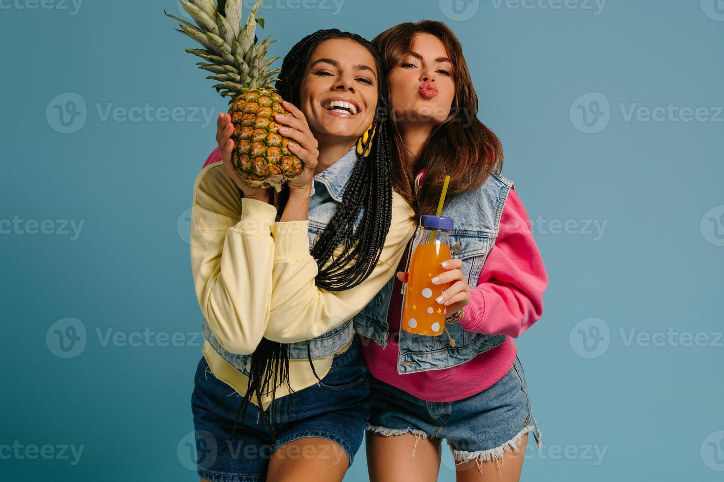 Two cheerful young women holding pineapple and bottle with lemonade against blue background photo