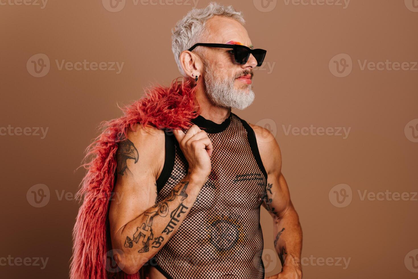 Fashionable mature gay man in transparent shirt carrying fluffy coat on shoulder against brown background photo