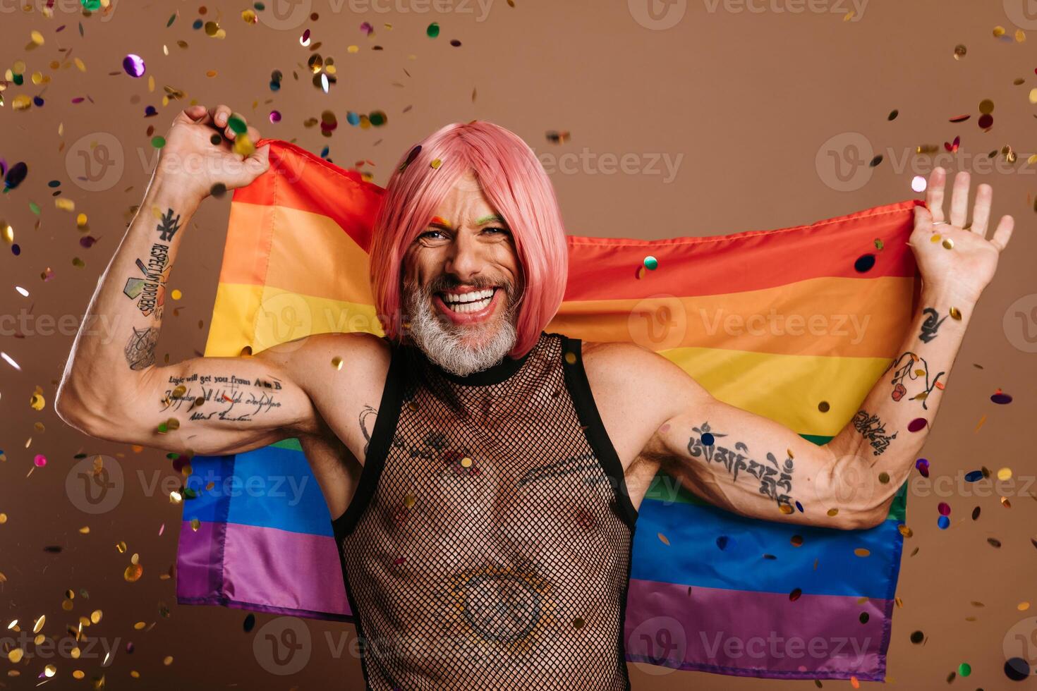 Joyful gay man in pink wig carrying rainbow flag while confetti flying around him against brown background photo