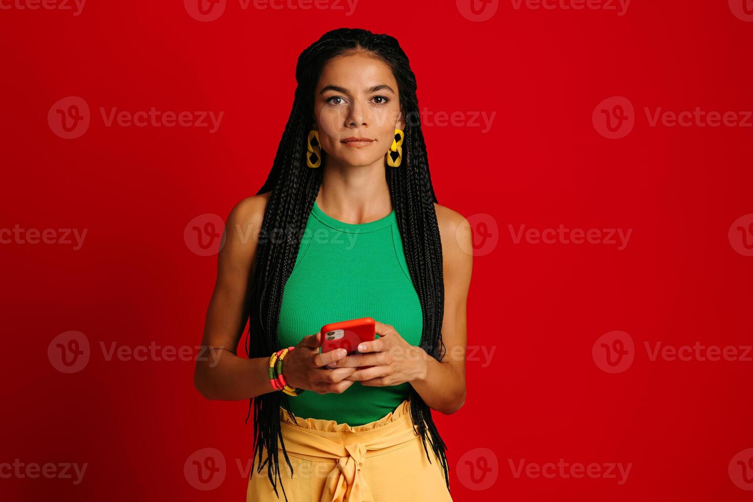 Attractive young Hispanic woman looking at camera while holding smart phone against red background photo