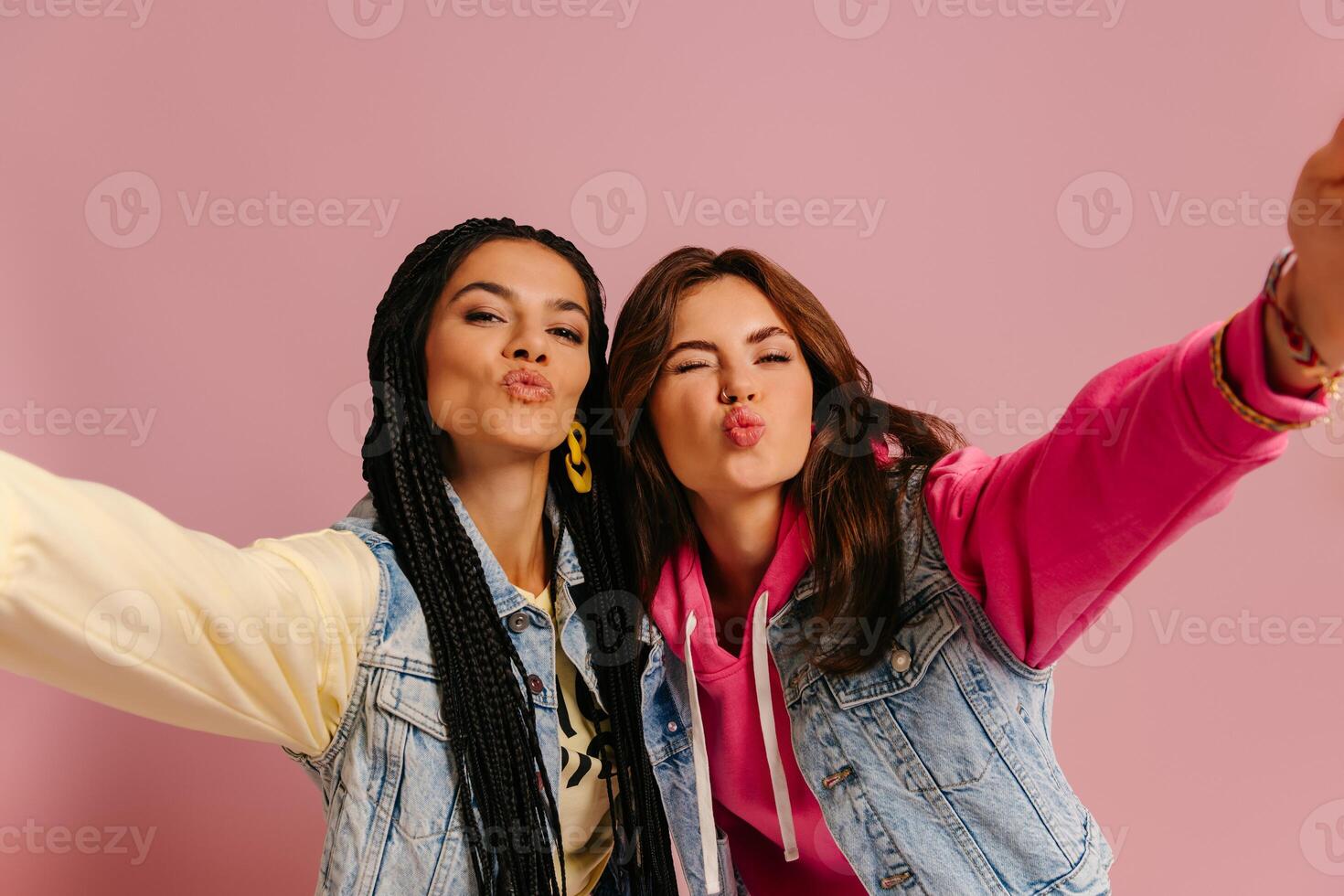 Two playful young women in colorful wear embracing and making self portrait against pink background photo