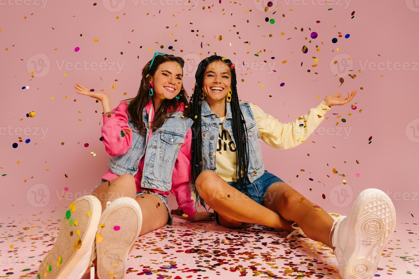 Two attractive young women throwing colorful confetti and smiling while sitting against pink background photo