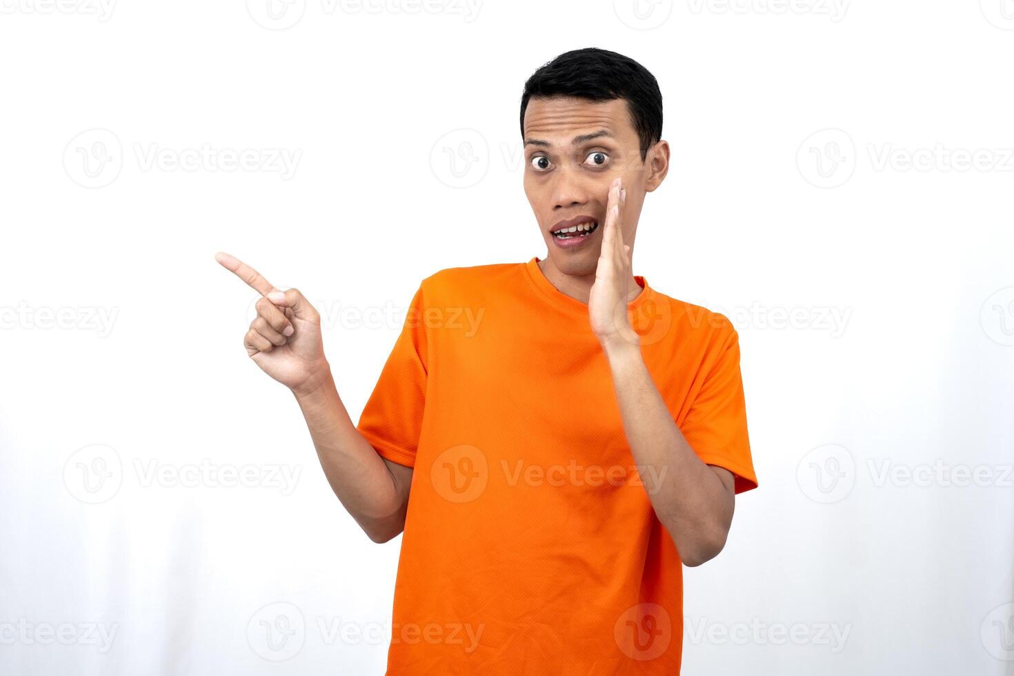 Portrait of a young Asian man wearing an orange t-shirt with his hand near his mouth in a whispering pose while pointing to the side of the copy space. Isolated on white background. photo