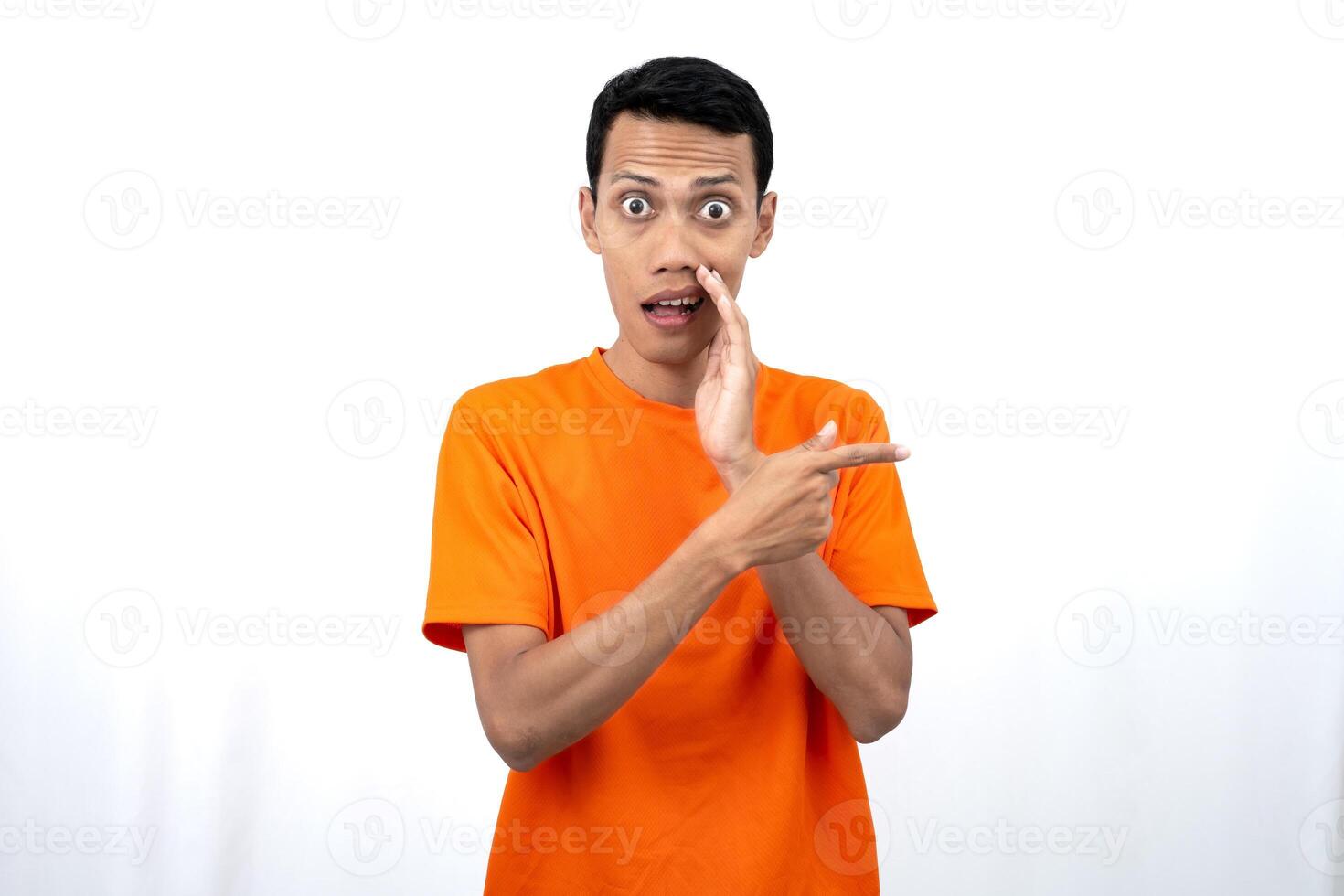 Portrait of a young Asian man wearing an orange t-shirt with his hand near his mouth in a whispering pose while pointing to the side of the copy space. Isolated on white background. photo