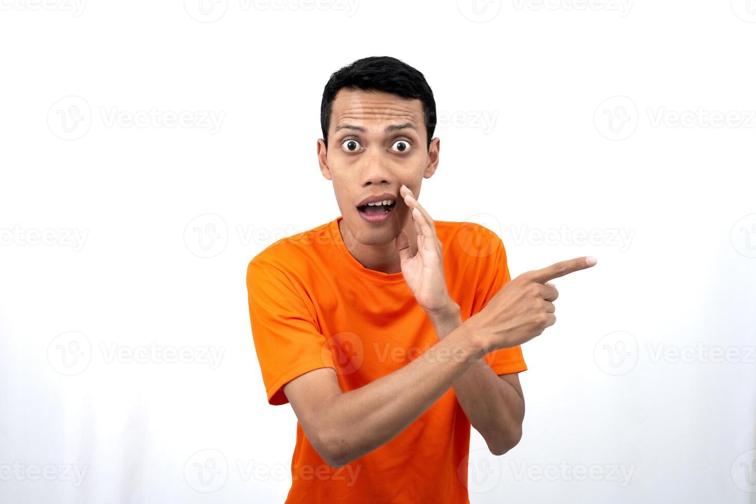 Portrait of a young Asian man wearing an orange t-shirt with his hand near his mouth in a whispering pose while pointing to the side of the copy space. Isolated on white background. photo
