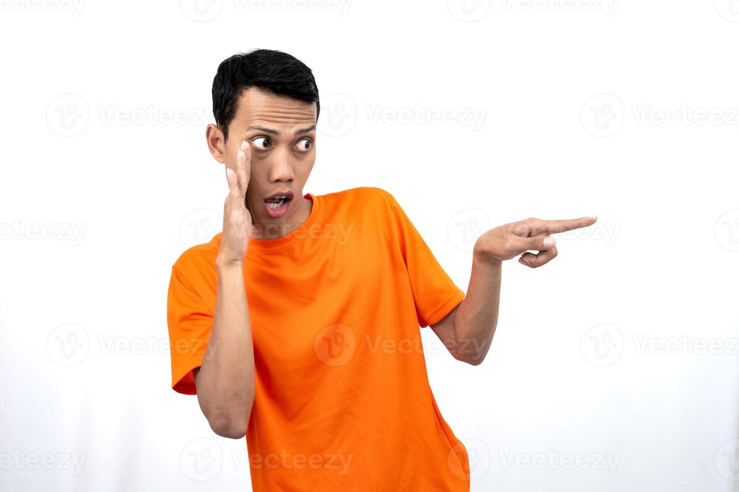 Portrait of a young Asian man wearing an orange t-shirt with his hand near his mouth in a whispering pose while pointing to the side of the copy space. Isolated on white background. photo
