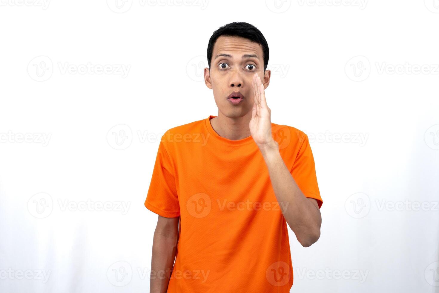 Portrait of a young Asian man wearing an orange t-shirt with his hand near his mouth whispering gossip and telling secrets while sharing news isolated on white background. photo