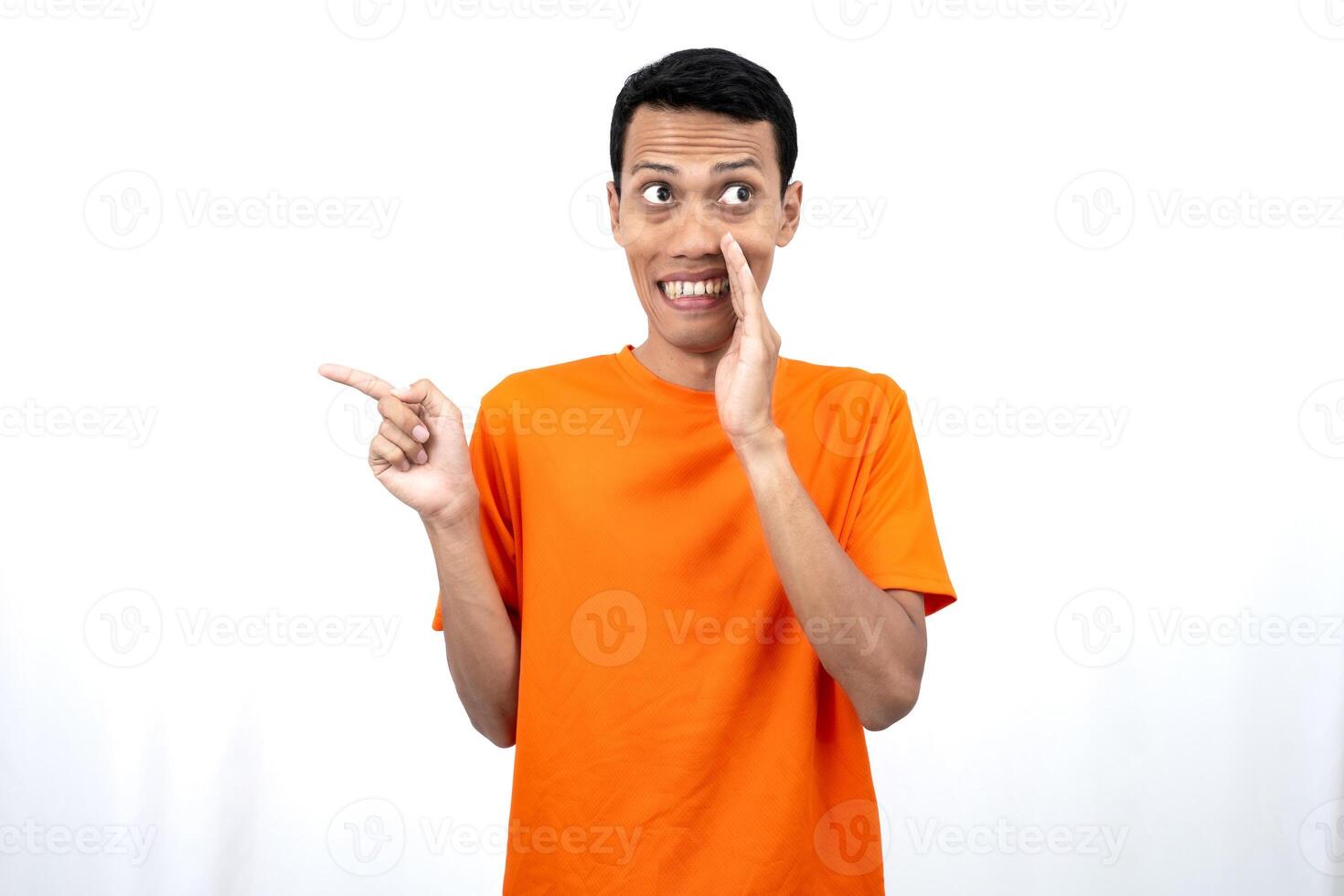 Portrait of a young Asian man wearing an orange t-shirt with his hand near his mouth in a whispering pose while pointing to the side of the copy space. Isolated on white background. photo