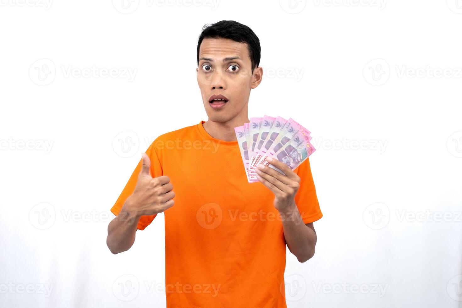 Portrait of a young Asian man wearing an orange t-shirt with a happy, surprised, pleased expression while carrying hundreds of thousands of rupiah. isolated on a white background. photo