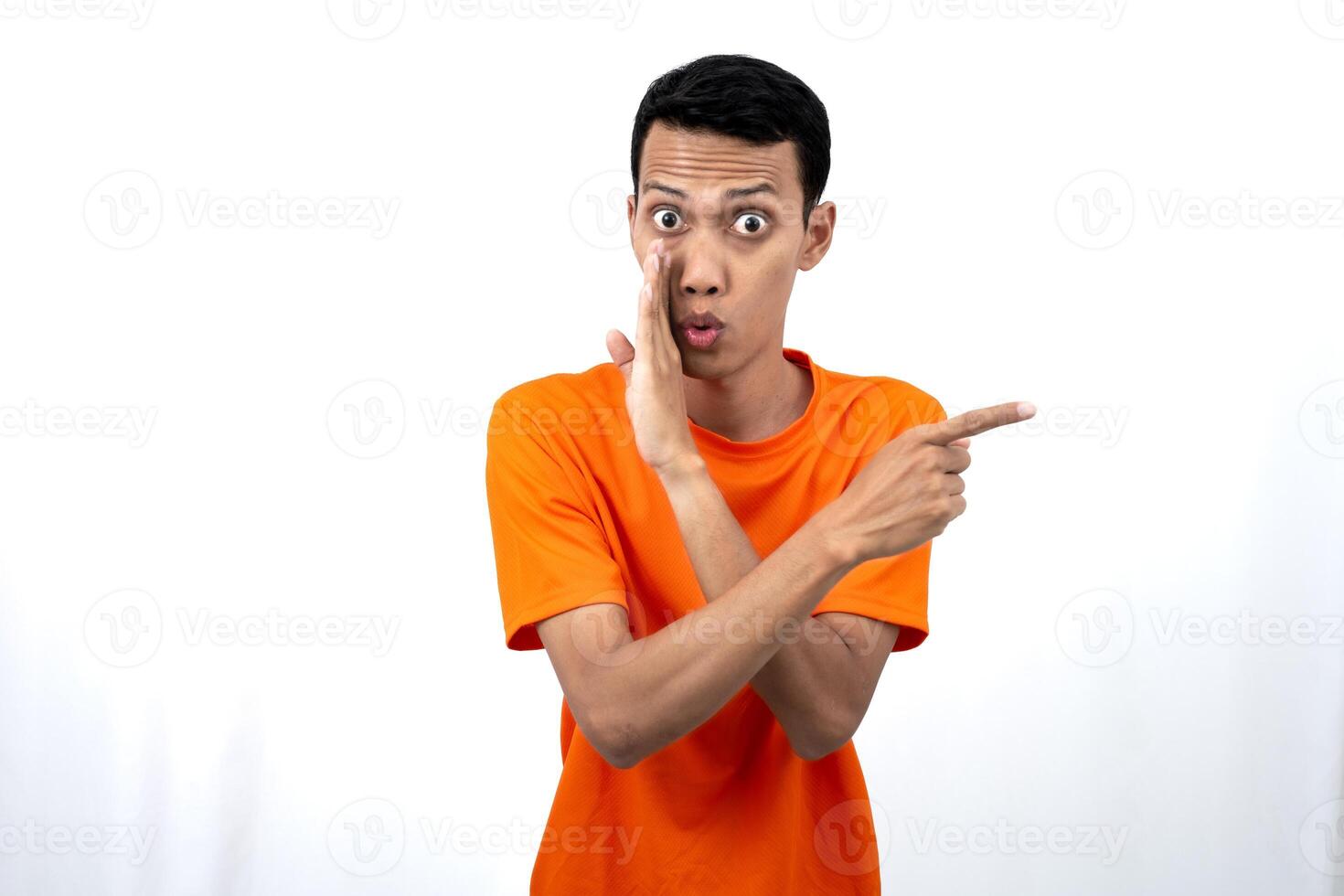 Portrait of a young Asian man wearing an orange t-shirt with his hand near his mouth in a whispering pose while pointing to the side of the copy space. Isolated on white background. photo