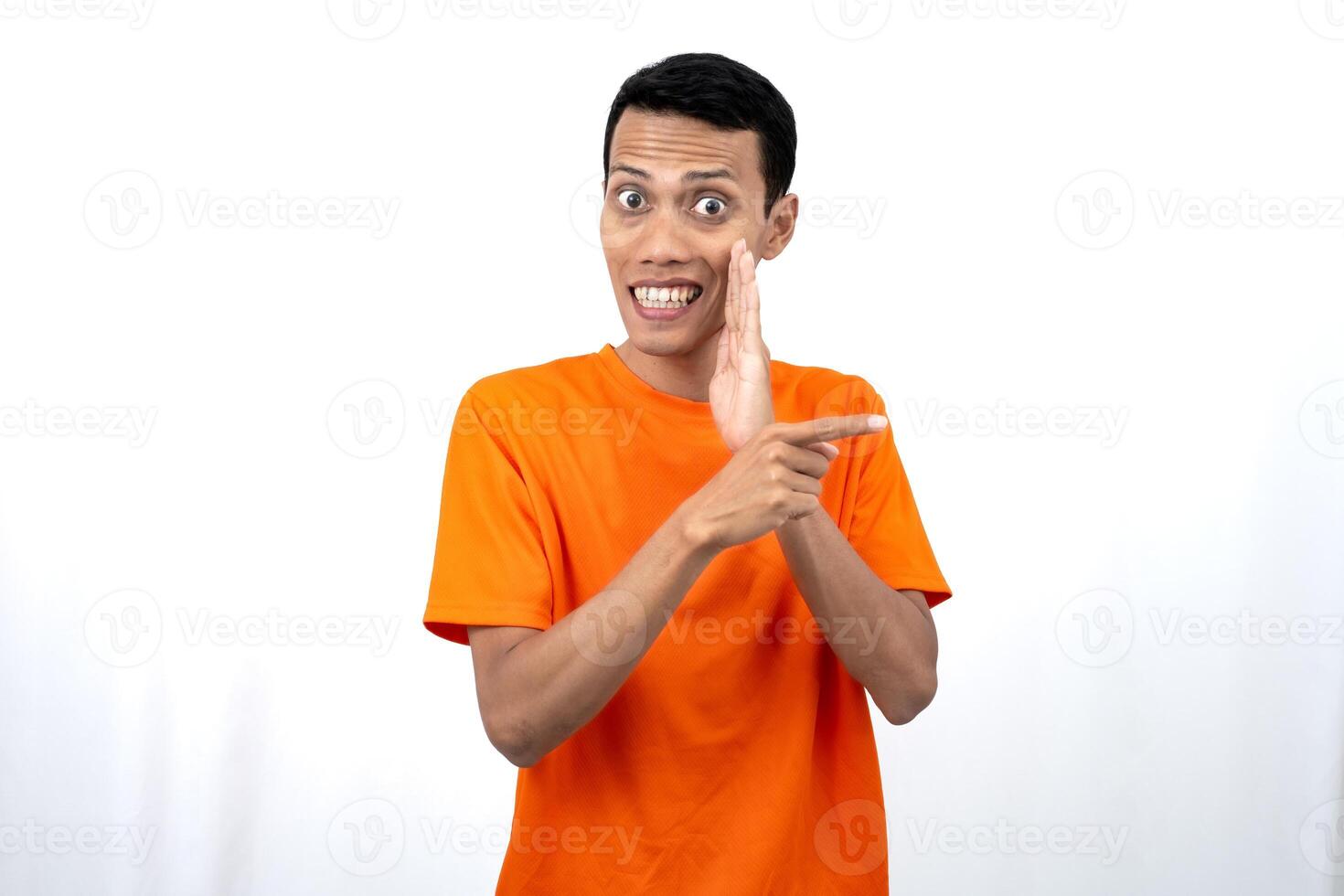 Portrait of a young Asian man wearing an orange t-shirt with his hand near his mouth in a whispering pose while pointing to the side of the copy space. Isolated on white background. photo