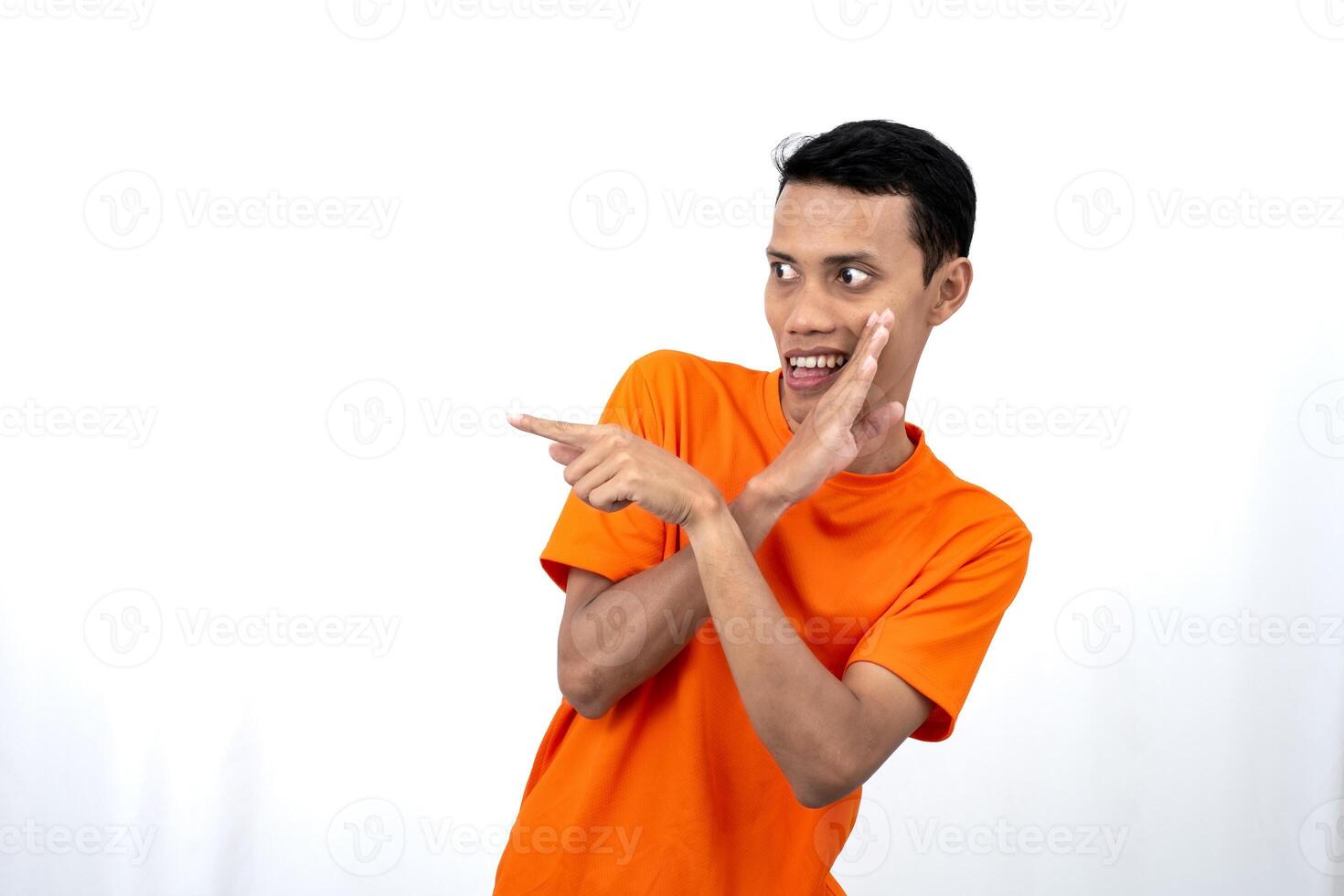 Portrait of a young Asian man wearing an orange t-shirt with his hand near his mouth in a whispering pose while pointing to the side of the copy space. Isolated on white background. photo
