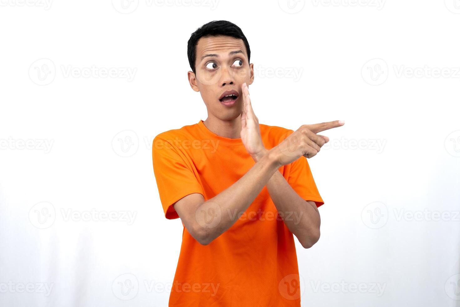Portrait of a young Asian man wearing an orange t-shirt with his hand near his mouth in a whispering pose while pointing to the side of the copy space. Isolated on white background. photo