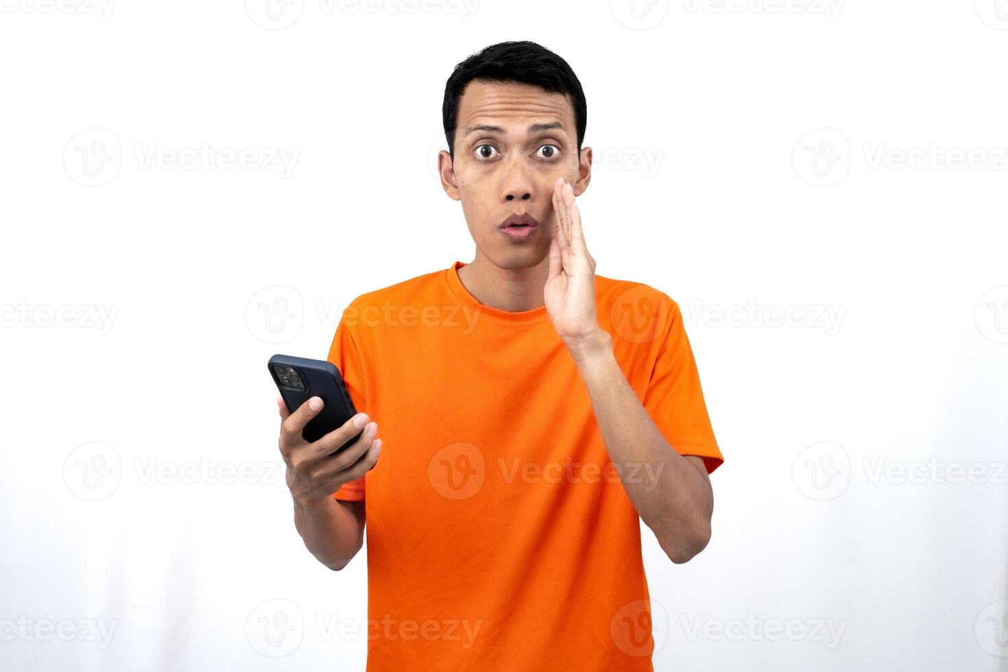 Portrait of a young Asian man wearing an orange t-shirt with his hand near his mouth whispering gossip and telling secrets while holding a smartphone. Sharing news isolated on white background. photo