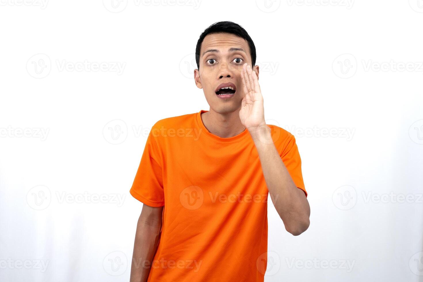 Portrait of a young Asian man wearing an orange t-shirt with his hand near his mouth whispering gossip and telling secrets while sharing news isolated on white background. photo