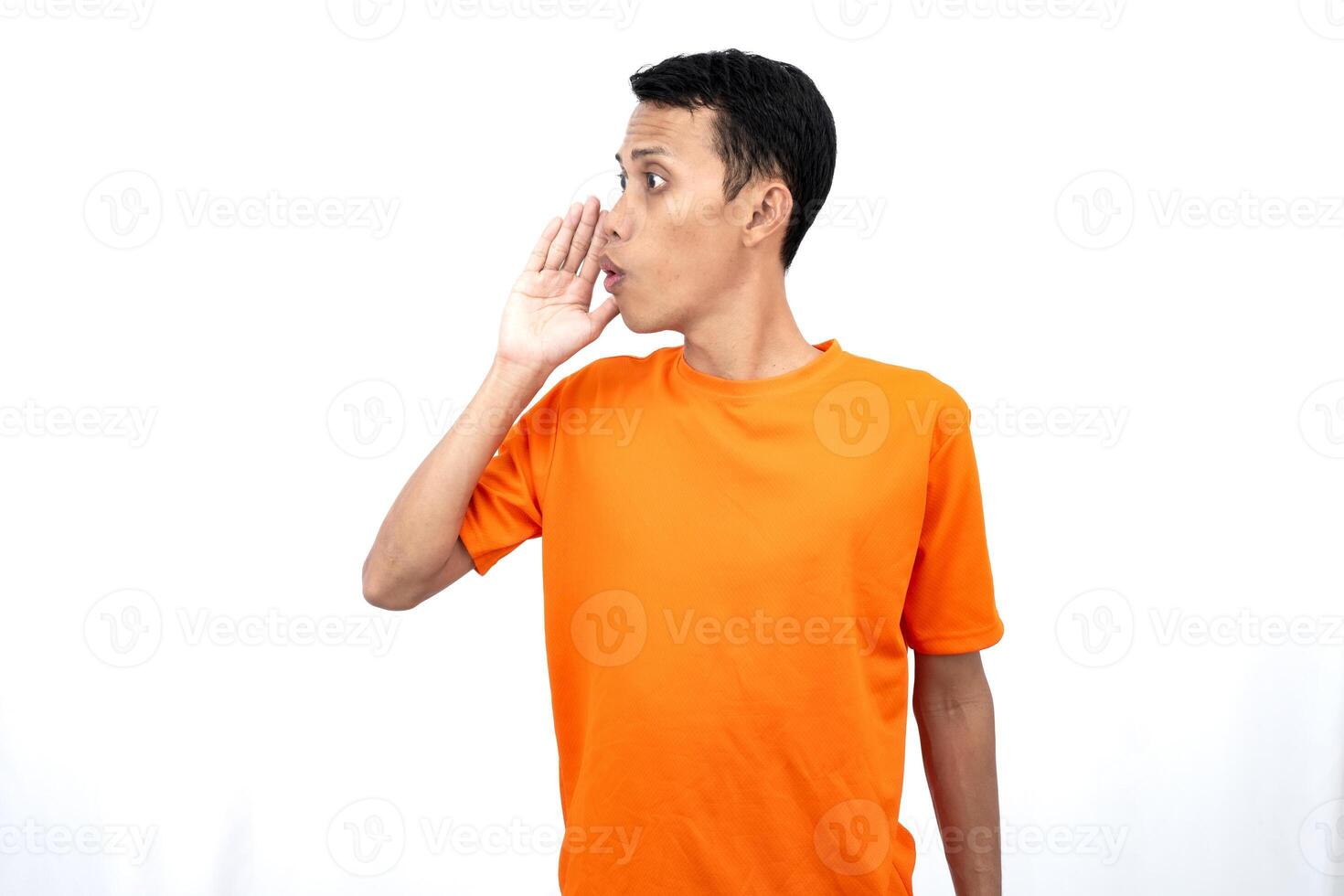 Portrait of a young Asian man wearing an orange t-shirt with his hand near his mouth whispering gossip and telling secrets while sharing news isolated on white background. photo