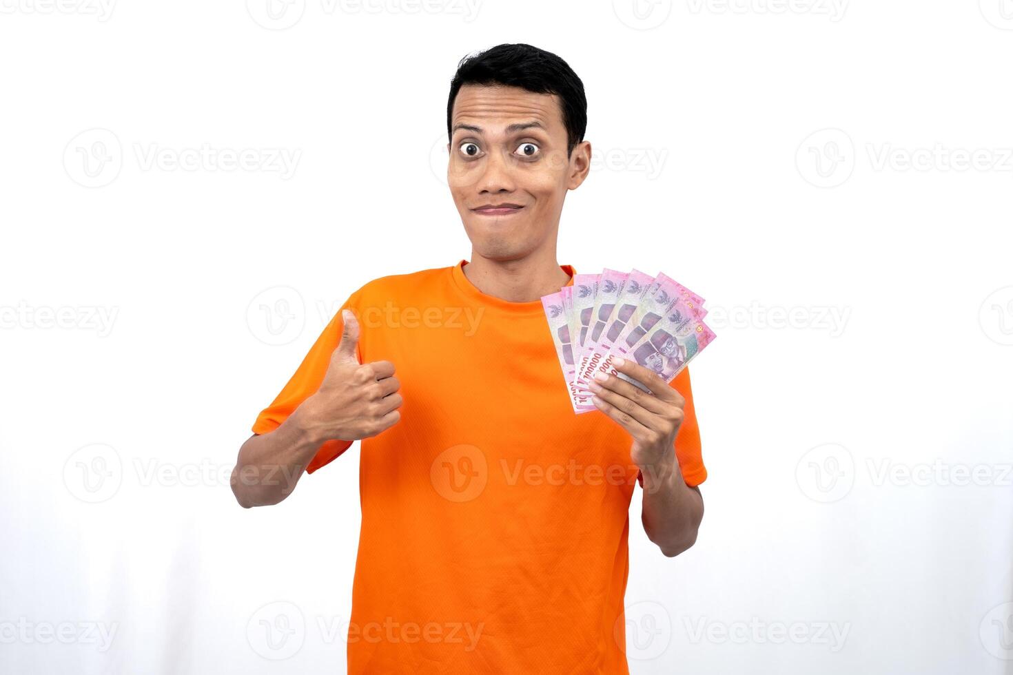 Portrait of a young Asian man wearing an orange t-shirt with a happy, surprised, pleased expression while carrying hundreds of thousands of rupiah. isolated on a white background. photo