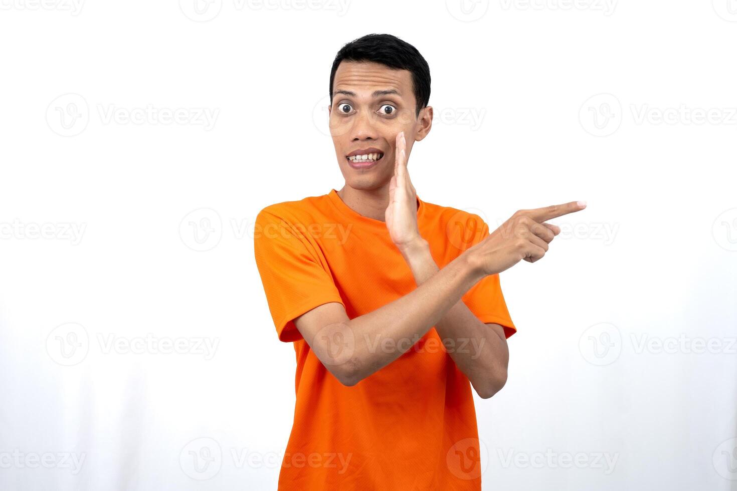Portrait of a young Asian man wearing an orange t-shirt with his hand near his mouth in a whispering pose while pointing to the side of the copy space. Isolated on white background. photo