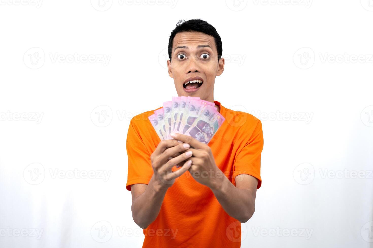 Portrait of a young Asian man wearing an orange t-shirt with a happy, surprised, pleased expression while carrying hundreds of thousands of rupiah. isolated on a white background. photo
