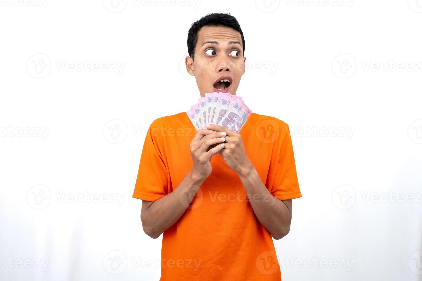 Portrait of a young Asian man wearing an orange t-shirt with a happy, surprised, pleased expression while carrying hundreds of thousands of rupiah. isolated on a white background. photo