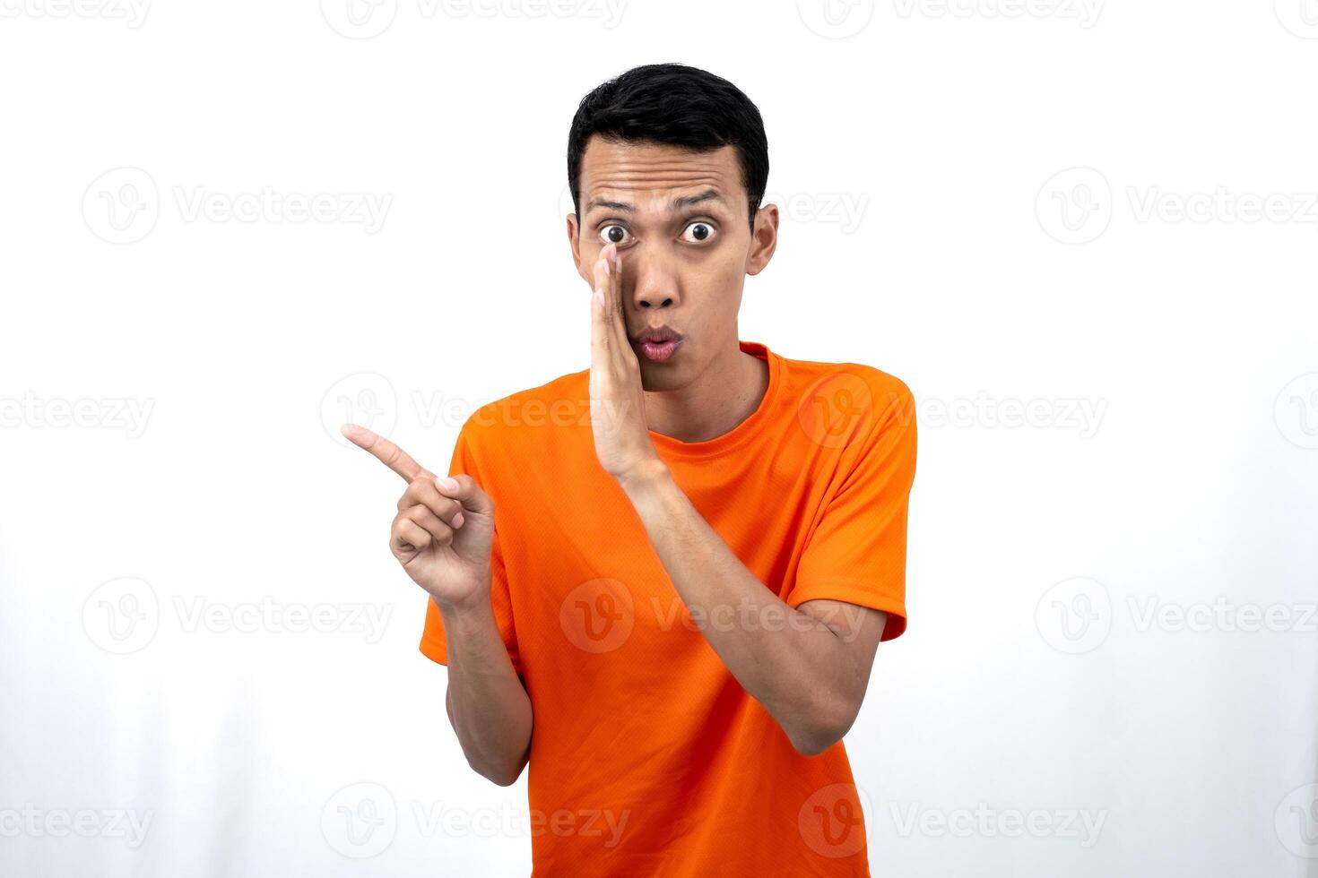 Portrait of a young Asian man wearing an orange t-shirt with his hand near his mouth in a whispering pose while pointing to the side of the copy space. Isolated on white background. photo
