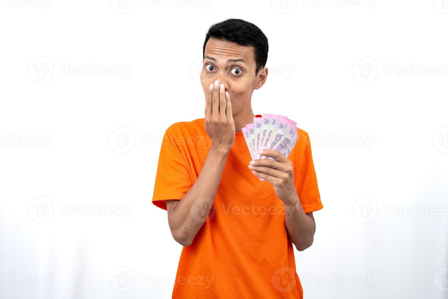 Portrait of a young Asian man wearing an orange t-shirt with a happy, surprised, pleased expression while carrying hundreds of thousands of rupiah. isolated on a white background. photo