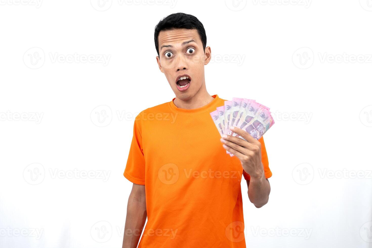 Portrait of a young Asian man wearing an orange t-shirt with a happy, surprised, pleased expression while carrying hundreds of thousands of rupiah. isolated on a white background. photo
